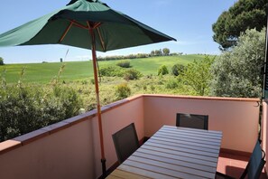 Un balcon avec une table et des chaises, un parasol vert et une vue sur un champ verdoyant et des arbres.
