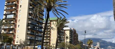 Un edificio residencial de varios pisos con balcones, palmeras y una acera.