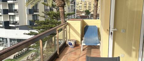 Un balcón con una mesa y sillas de vidrio, una puerta, y vista a un edificio residencial con balcones y palmeras.