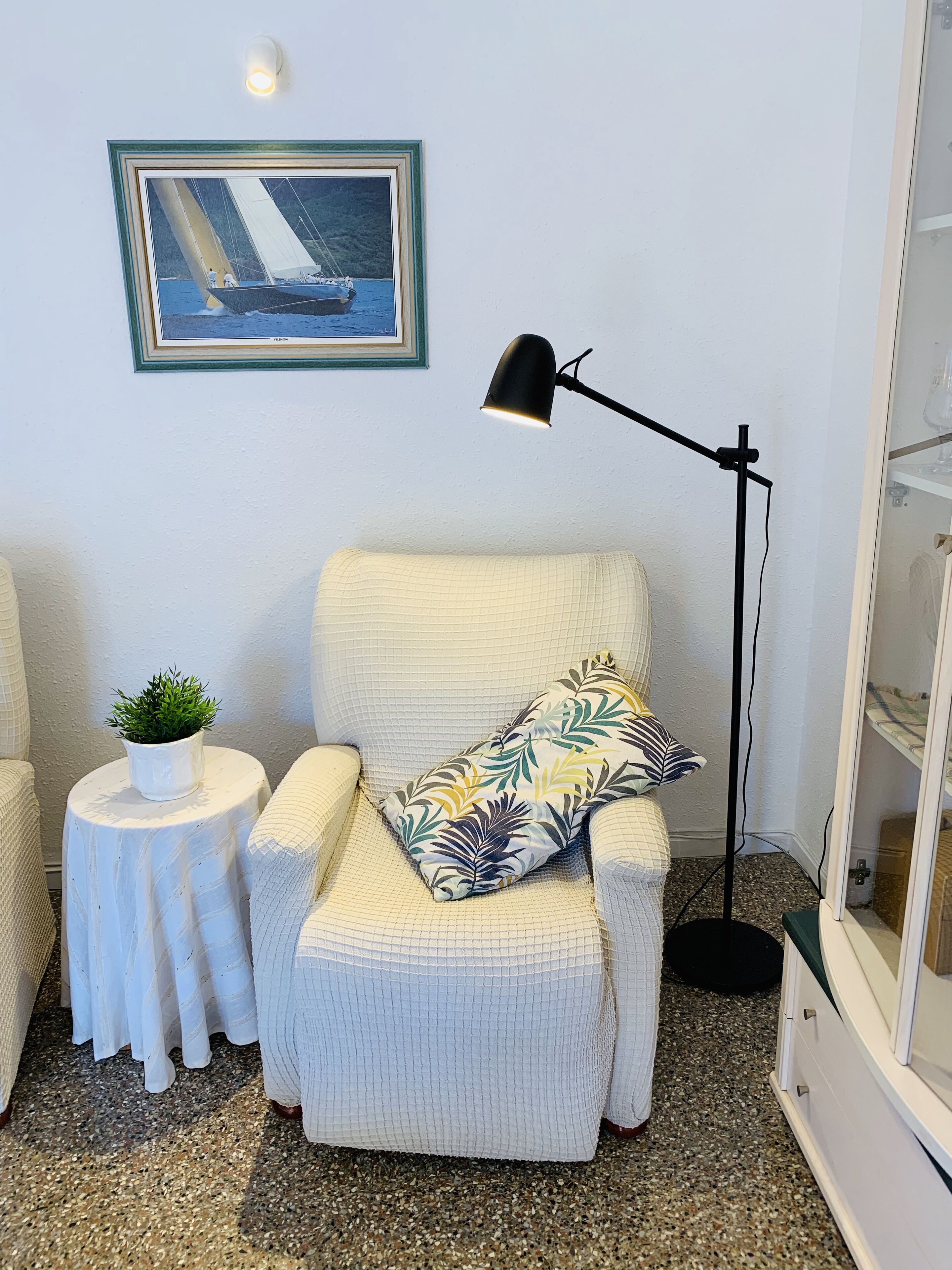 A cozy room with a white armchair, a small round table with a plant, a framed painting of a sailboat, and a floor lamp.