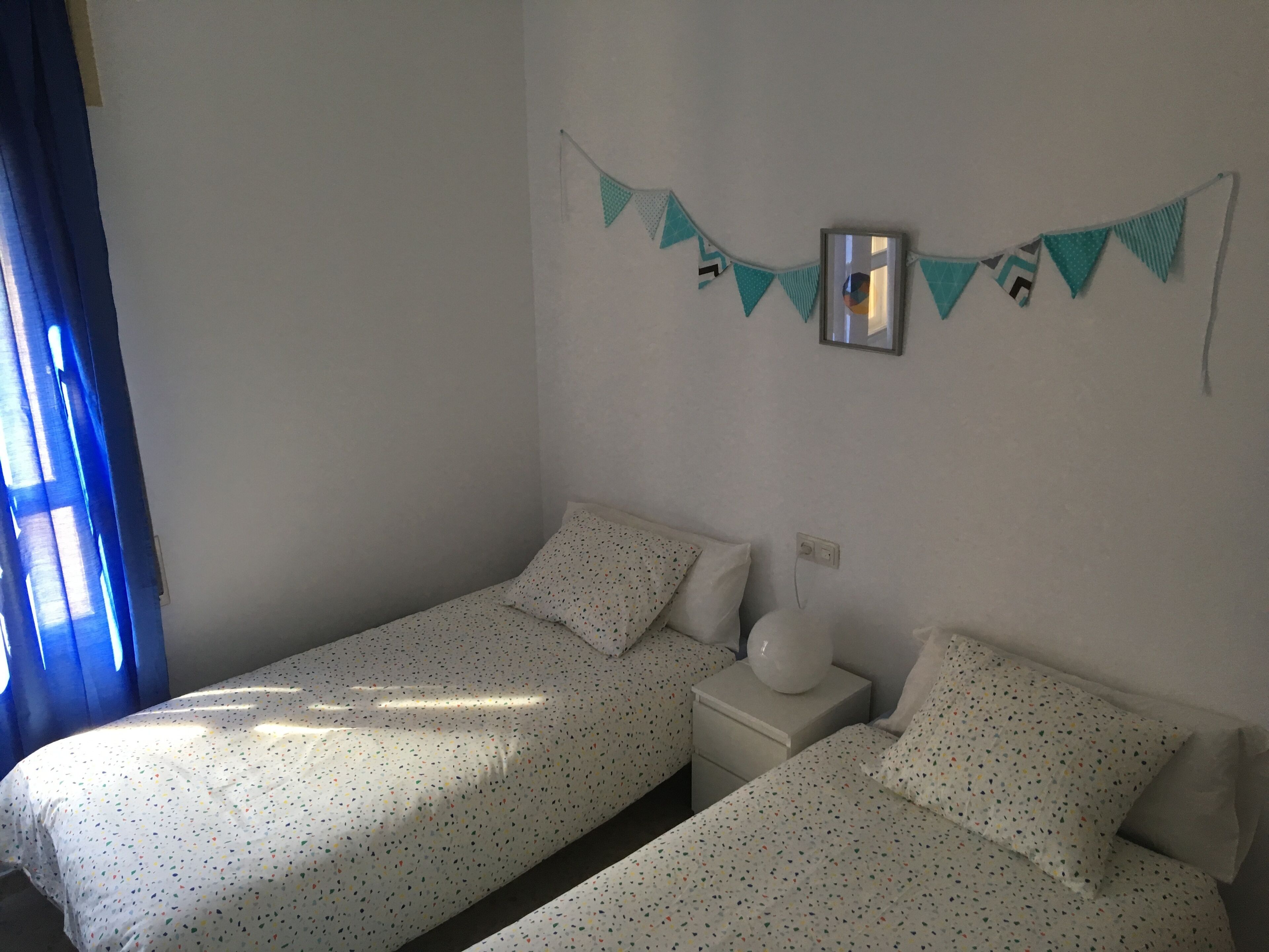 Una camera da letto con due letti singoli, un comodino con una lampada, uno specchio fissato alla parete e una ghirlanda decorativa color acquamarina.
