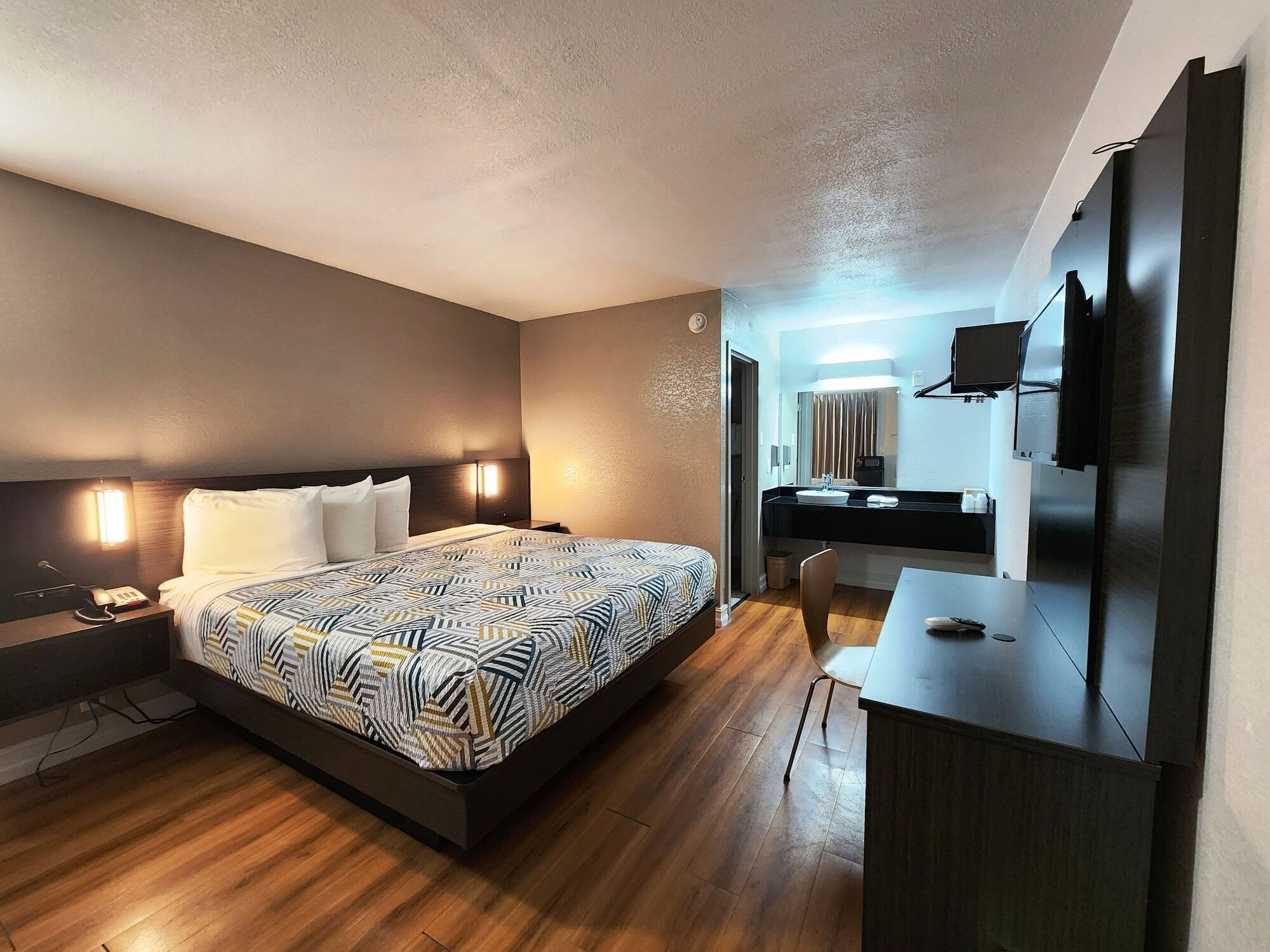 A modern hotel room with a large bed, bedside table with a lamp, a desk with a chair, a flat-screen TV mounted on the wall, and a bathroom visible through an open door.