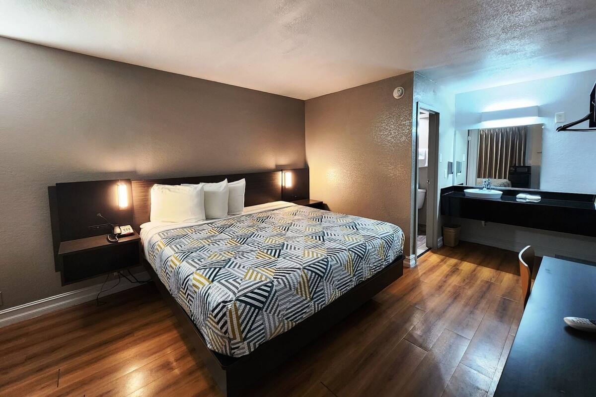 A hotel room with a large bed, bedside table, lamp, and a small desk with a chair.