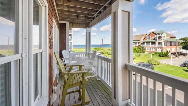 En träveranda med vita räcken och stolar, med utsikt över en strand och ett stort hus.