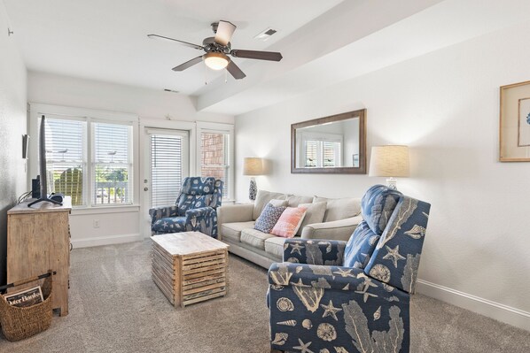 A living room with a ceiling fan, a sofa, a chair, a coffee table, and a window with blinds. - Sunset Sound: Water Views, Shared Pool, Elevator (Nags Head)