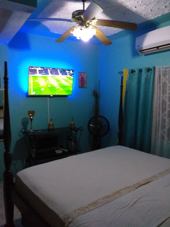 A bedroom with a bed, a ceiling fan, a TV mounted on the wall, and a window with curtains.