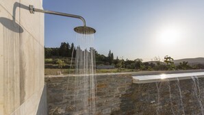 A modern outdoor shower with water cascading down a stone wall, set against a backdrop of trees and a clear sky. - Virginia Luxury Villas (Samos)