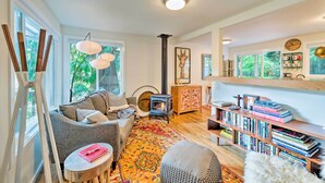 A cozy living room with a fireplace, a bookshelf, and a large window with a view of greenery. - House w/ Deck on Whidbey Island, 1 Mi from Shore! (Clinton)