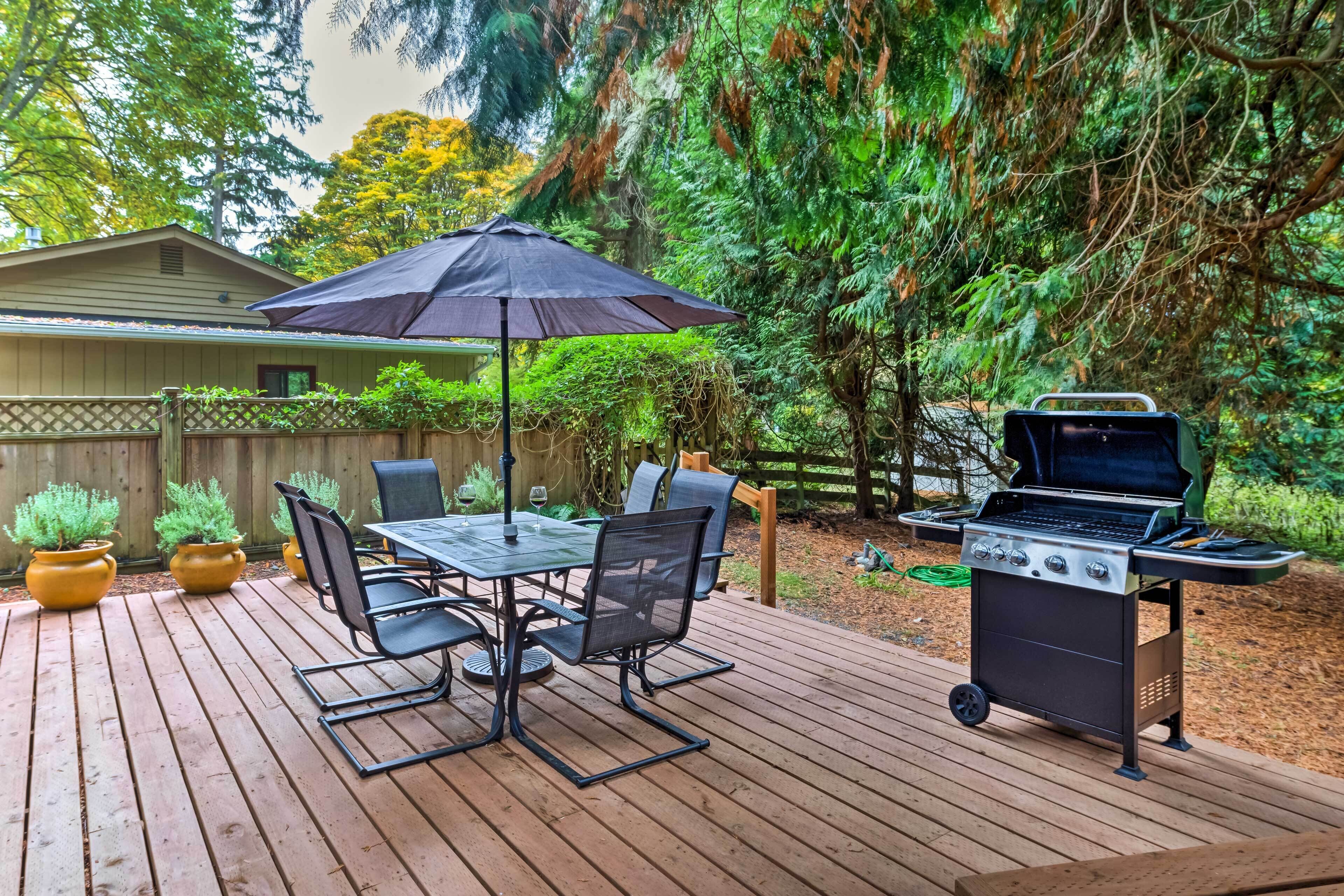 Una terraza de madera con una mesa, sillas, una parrilla y plantas en macetas.