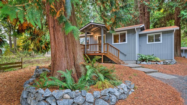 A cabin with a wooden deck, stone wall, and a large tree in the foreground. - House w/ Deck on Whidbey Island, 1 Mi from Shore! (Clinton)