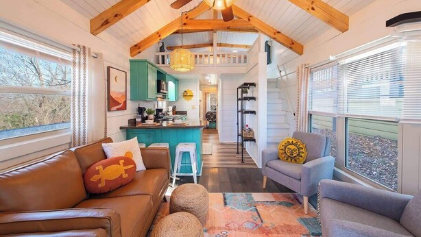 Un interior acogedor con sofá, sillón y reposapiés, vigas de madera, una cocina y vistas amplias a través de grandes ventanales.