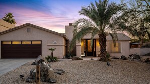 Una casa con garaje, entrada para vehículos y jardín ajardinado con rocas y una palmera.