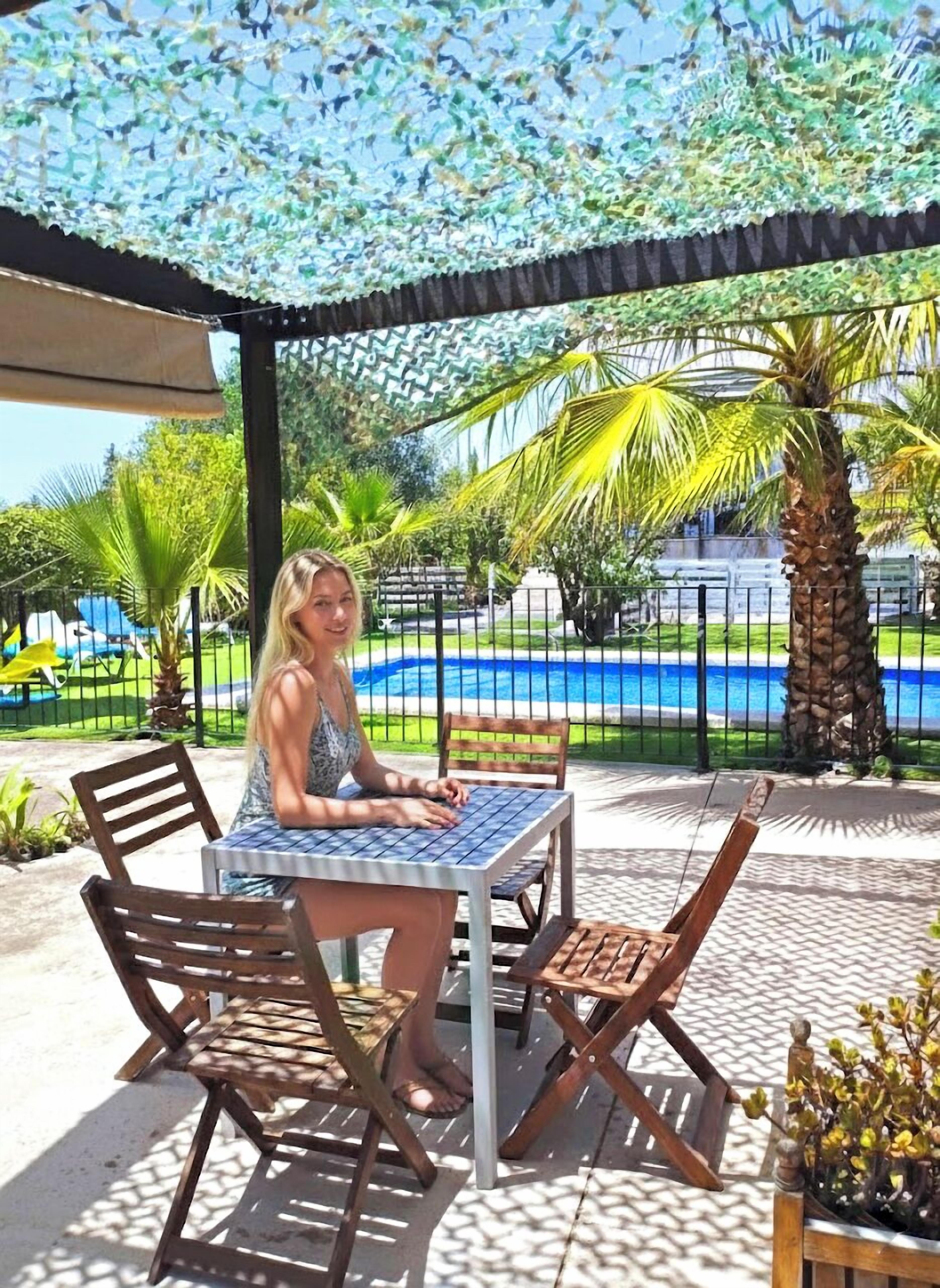 A person seated at a table with a view of a swimming pool and palm trees.