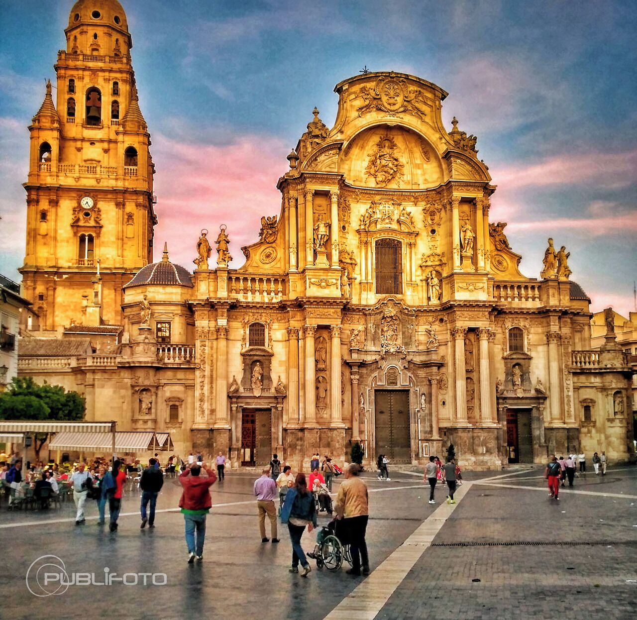 A historic building with a prominent bell tower and ornate facade, surrounded by a plaza with people and a wheelchair.