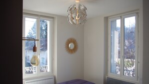Un dormitorio con cama, dos ventanas, radiador, espejo en la pared y lámpara en el techo.
