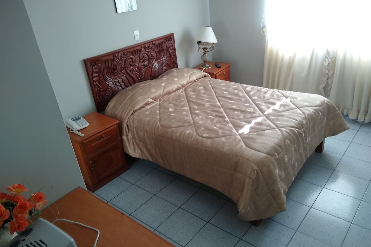 En snyggt bäddad säng med ett mönstrat överkast, ett träbord vid sängen med en lampa, en telefon och ett litet bord med en projektor.
