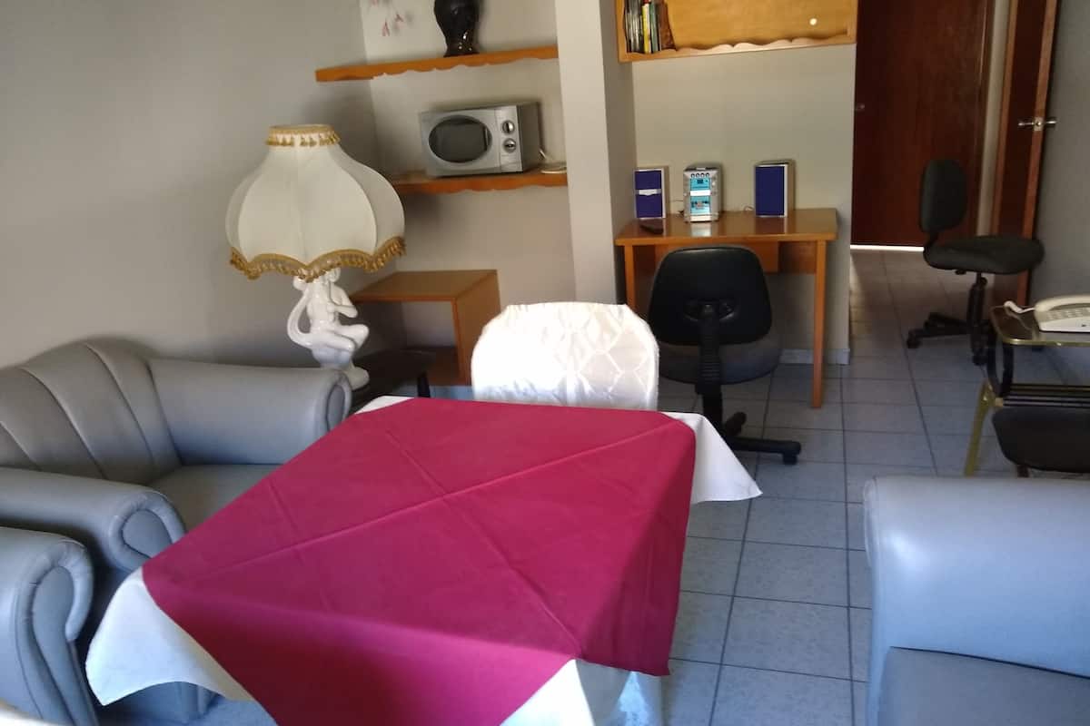 Ett rum med en säng täckt av ett rött överkast, en grå soffa, en vit lampa med en guldfärgad fot, en trähylla med böcker och en mikrovågsugn samt ett skrivbord med en dator och en telefon.