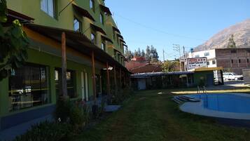 Un hotel con piscina, un área exterior techada y una montaña al fondo.