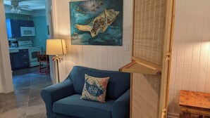 A living room with a blue sofa, a red ottoman, a painting of a sea turtle, a lamp, and a kitchen in the background. - Perfect Couples Getaway - Canal Front Apartment  (Eastpoint)