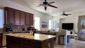 A modern kitchen with granite countertops, wooden cabinets, and a large island. There are ceiling fans, a dining area with a sofa, and a living room with a TV. - Luxurious Golf Course Views-Private Pool +2 King Suites-Sleeps 8 (San Tan Valley)