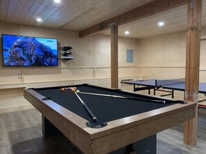 A pool table with a cue stick, a television displaying a nature scene, and ping pong tables in a room with wooden beams and a light-colored floor.