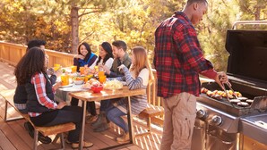 Un groupe de personnes est réuni autour d’une table en plein air, sur une terrasse en bois, près d’un barbecue.