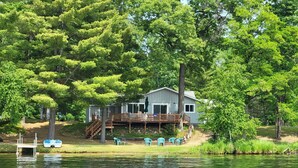 A lakeside cabin with a deck, surrounded by trees and a dock with boats. - LAKEFRONT COTTAGE  (Lake George)
