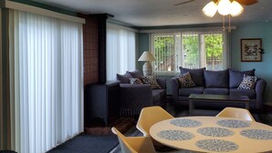 A living room with a ceiling fan, a round table with patterned centerpieces, a sofa with patterned cushions, and a window with blinds. - LAKEFRONT COTTAGE  (Lake George)