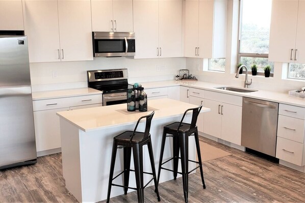 A modern kitchen with white cabinets, stainless steel appliances, a central island with bar stools, and a window providing natural light.