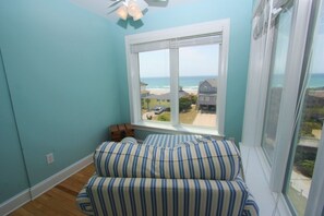 A room with a large window overlooking a beach, a striped sofa, and a ceiling fan. - Pool, Game Room, 5 Ensuite BRs & Ocean Views! (Emerald Isle)