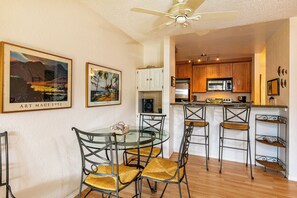 A dining area with a glass table and metal chairs, a kitchen with wooden cabinets and a microwave, and two framed artworks on the wall.