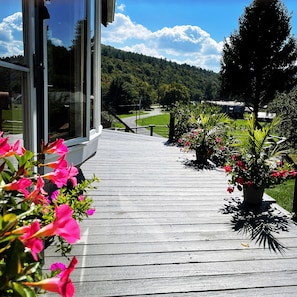 Une terrasse en bois menant à une maison dotée d’une porte en verre, entourée de plantes et de fleurs en pot.