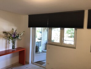 A room with a red bench, a vase with flowers, a window with blinds, and a view of the outside.