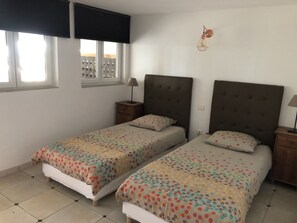 A bedroom with two beds, a window with blinds, a nightstand with a lamp, and a wall-mounted light fixture.