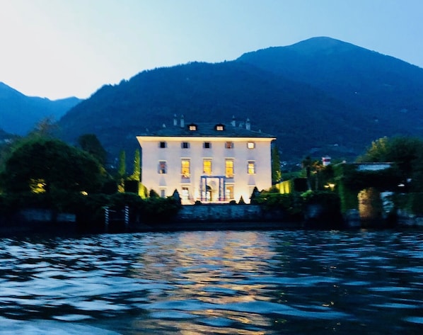 A large, white building with multiple windows and a symmetrical facade, situated by a body of water with reflections.