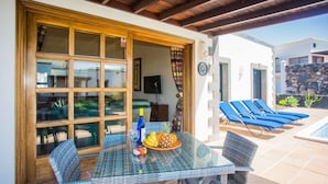 An outdoor dining area with a glass table and wicker chairs, a poolside with blue lounge chairs, and a stone wall in the background. - Villa Eleonor in Playa Blanca (Playa Blanca)