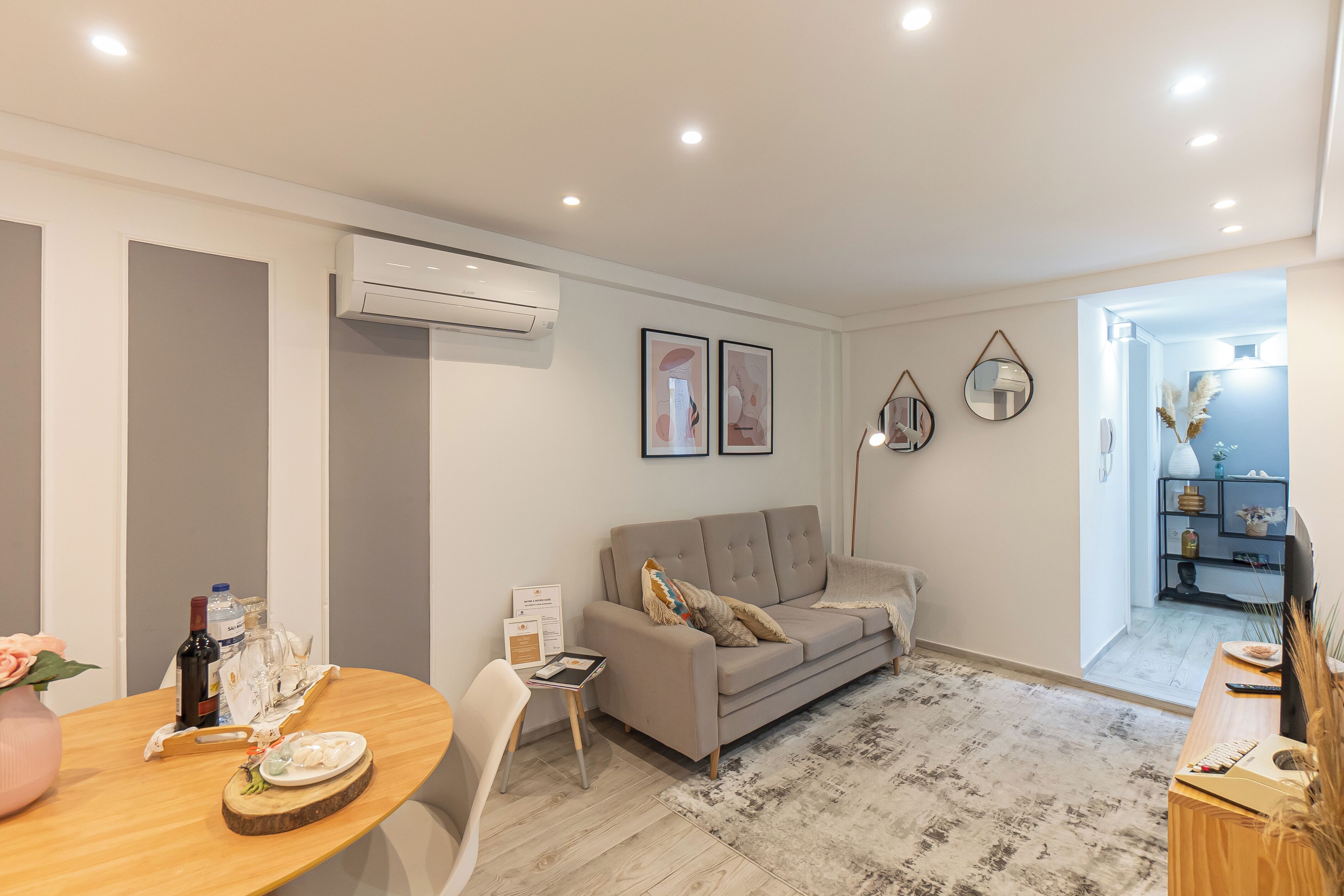 Una sala de estar moderna con sofá, una mesa de comedor y una mesa de centro. Hay un equipo de aire acondicionado en la pared y un cuadro enmarcado sobre el sofá.