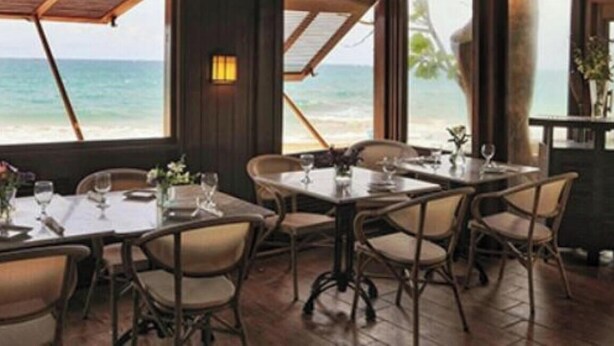 Un comedor con mesas y sillas, ventanas amplias con vista al océano y suelo de madera.