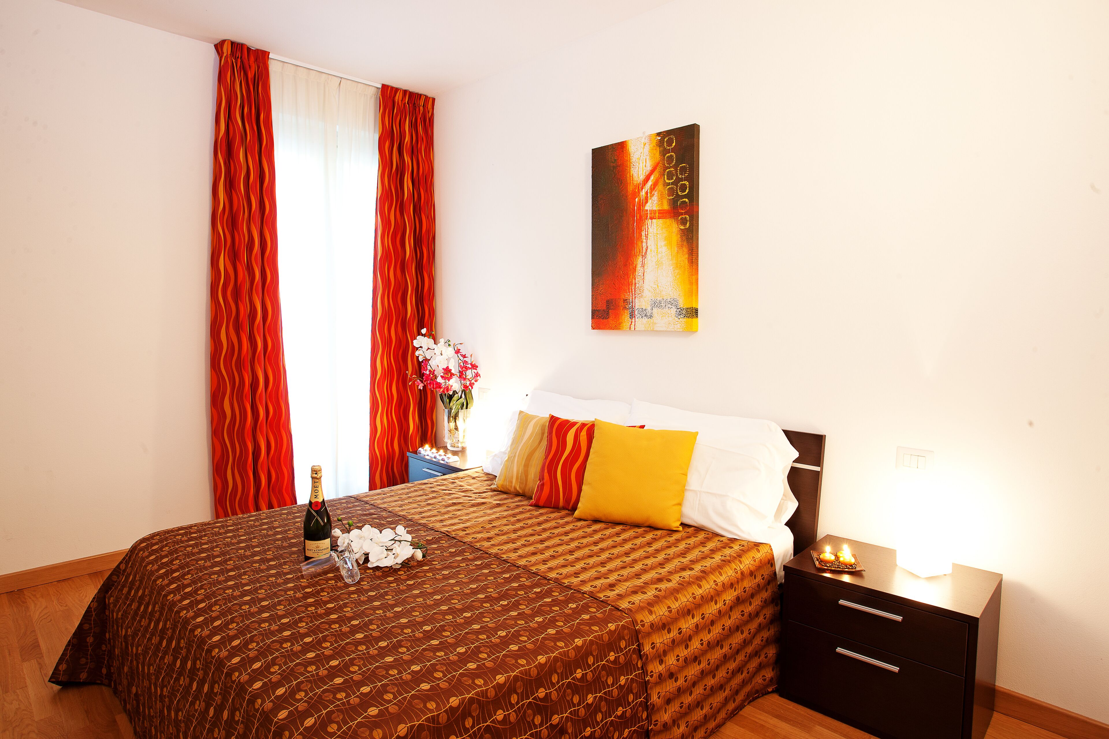 A hotel room with a bed, a nightstand, a bottle of champagne, and a painting on the wall.