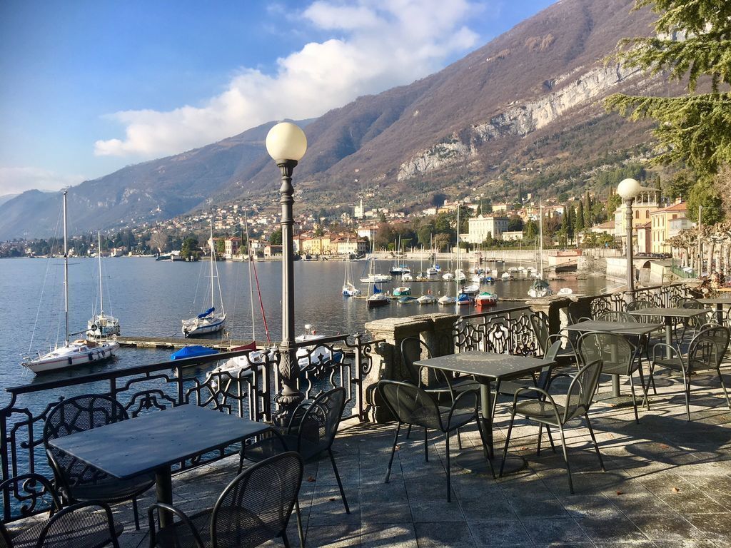 Um café à beira-mar com mesas ao ar livre, com vista para um porto e montanhas ao fundo.
