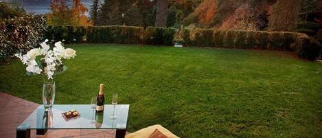 An outdoor dining area with a glass table, wine glasses, and a bottle of champagne, set against a backdrop of a grassy lawn and trees.