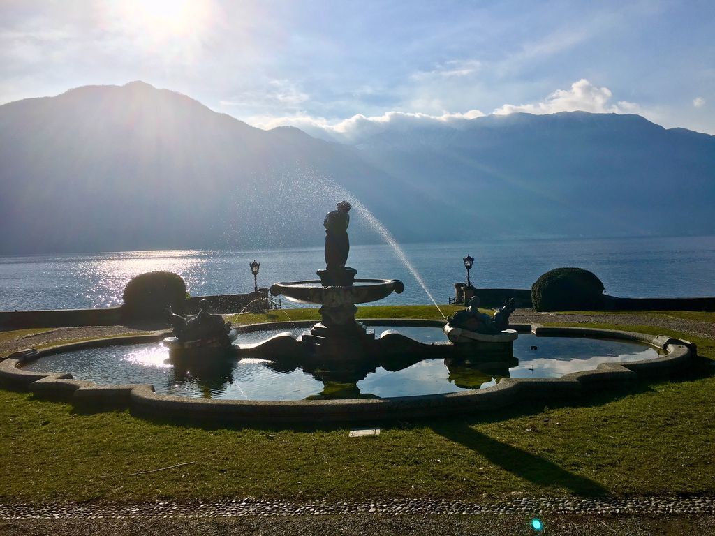 Uma fonte com uma estátua no centro, rodeada por um lago circular, e montanhas ao fundo.