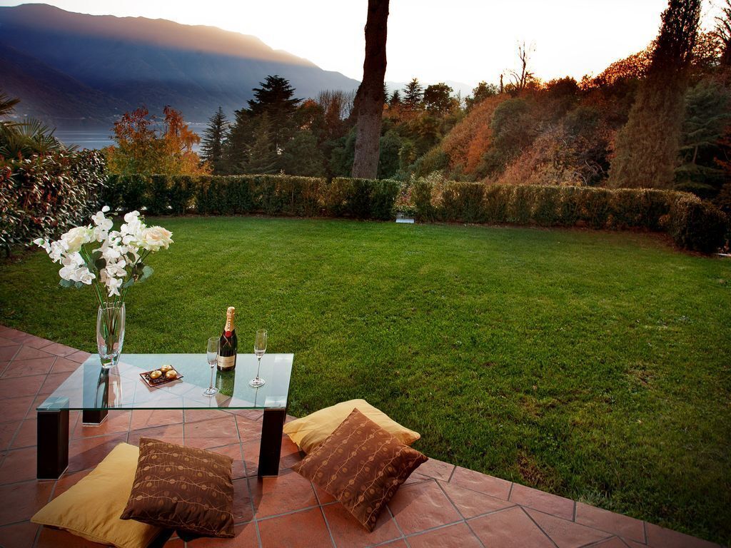 An outdoor dining area with a glass table, wine glasses, and a bottle of champagne, set against a backdrop of a grassy lawn and trees.