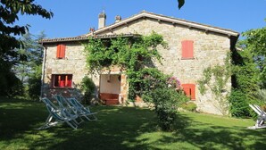 Una casa de pedra amb persianes vermelles, un jardí amb cadires verdes i un cel blau clar.