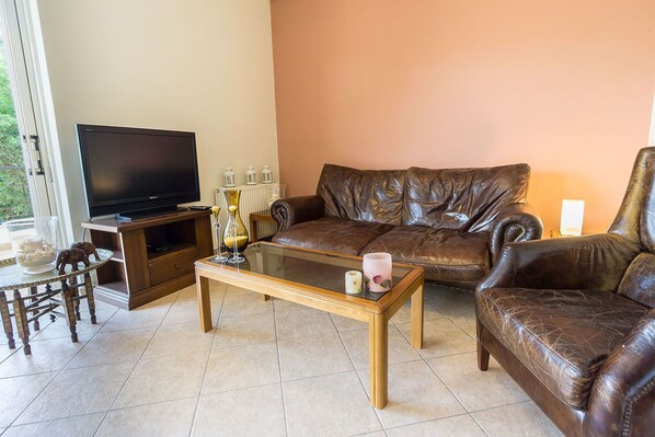A living room with a leather sofa, a glass coffee table, a television on a stand, and a small dining table with chairs.