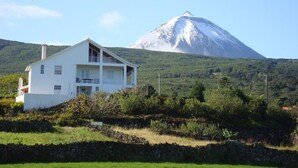 A white two-story house with a balcony, set against a backdrop of a snow-capped mountain and lush greenery. - PICO Hollidays  Rentals- Mountain and sea view for families or  groups (São Roque do Pico)