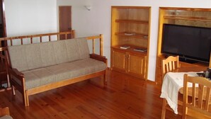 A wooden-floored living room with a sofa, a TV, and a dining area with chairs and a table. - PICO Hollidays  Rentals- Mountain and sea view for families or  groups (São Roque do Pico)