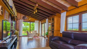 A cozy living room with a leather sofa, wooden flooring, a fireplace, and large windows offering a view of the outdoors.