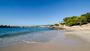 A beach with clear blue water, a sandy shore, and a white building with trees nearby.