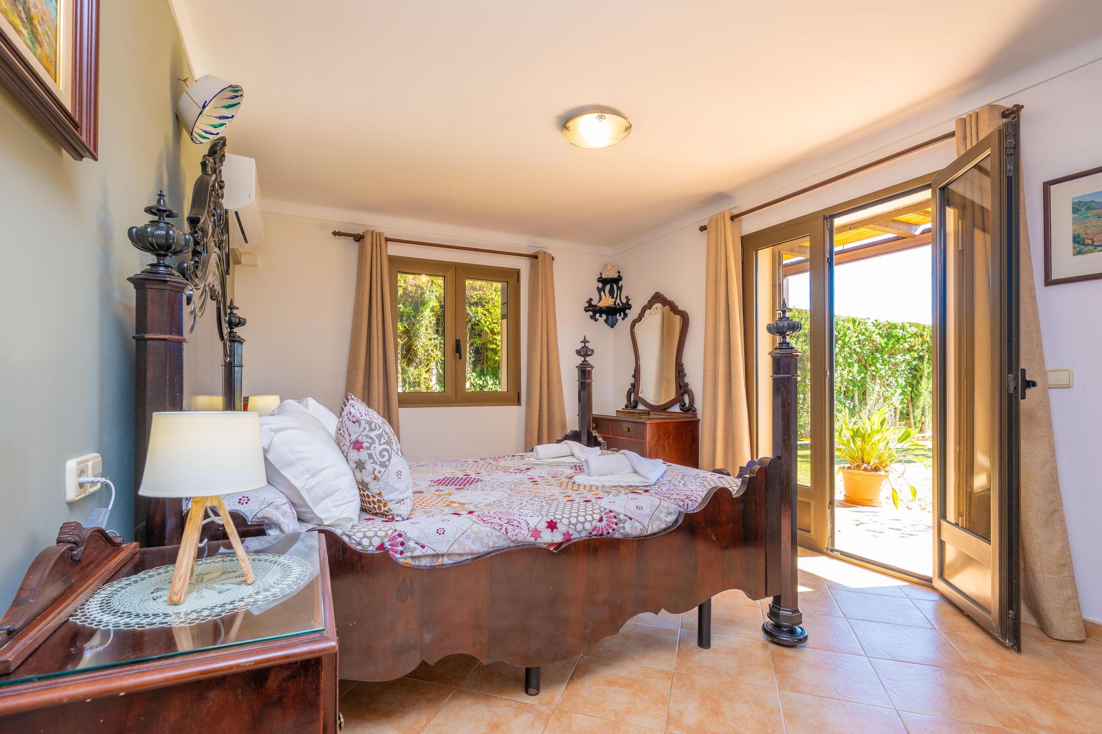 Une chambre avec un lit en bois, une table de chevet avec une lampe, une commode, une fenêtre avec des rideaux et une vue sur la verdure à l’extérieur.